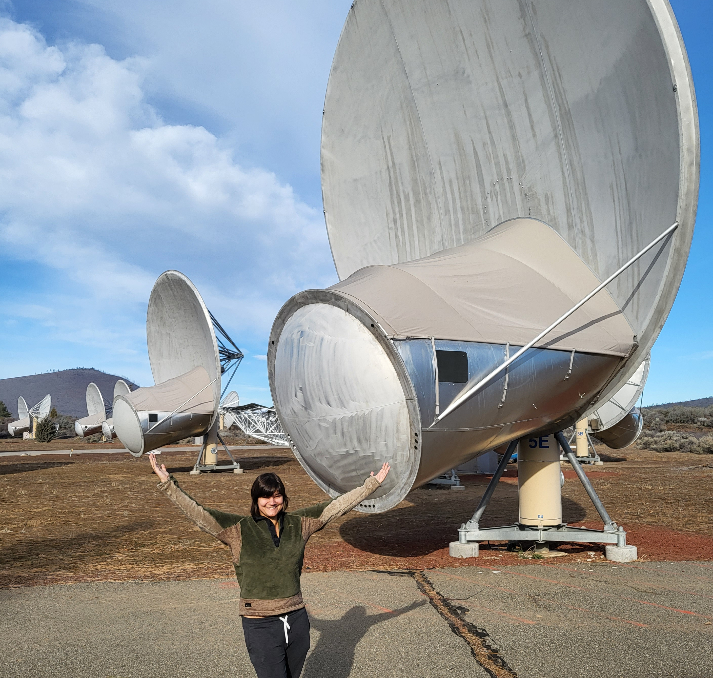 Dr. Sheikh stands in front of one of the dishes of the Allen Telescope Array with her arms up, imitating the dish. Other dishes are visible in the background